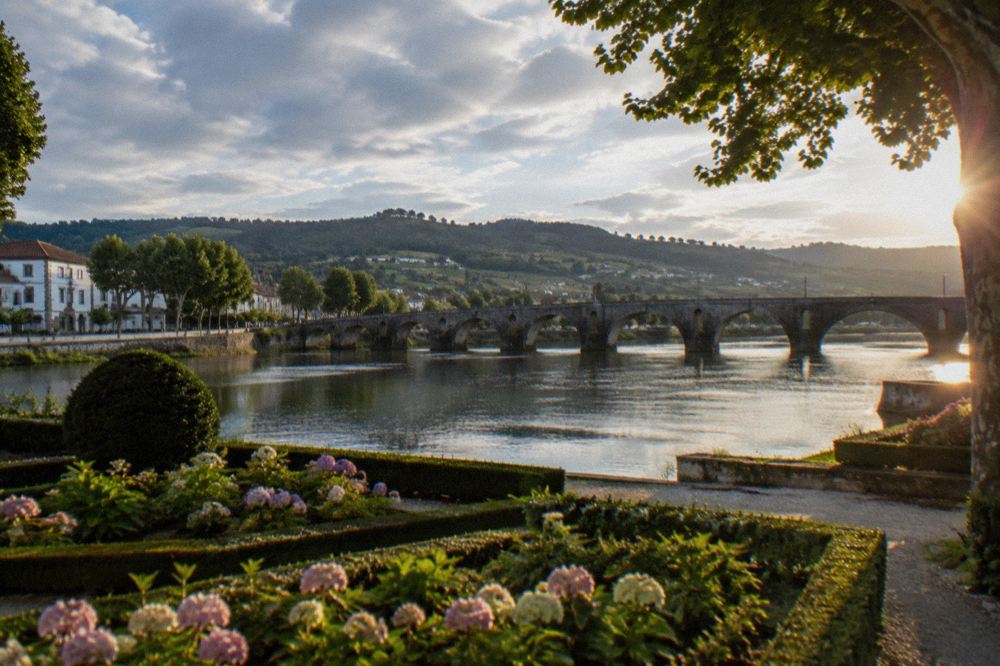 Flower beds and clipped hedges in a formal garden beside a river with a stone bridge in the distance