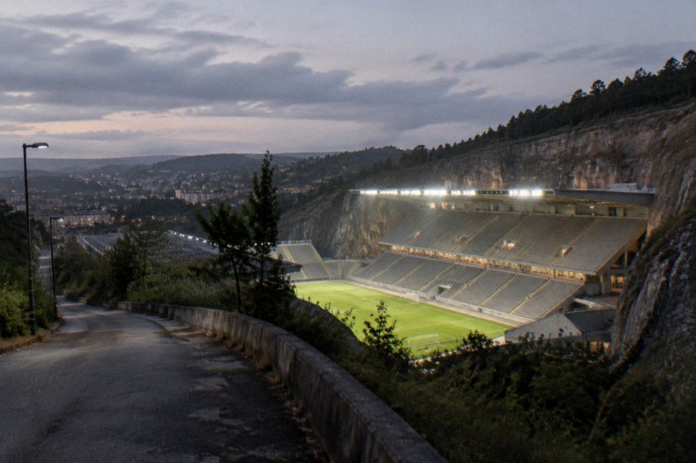 An evening view of a floodlit football stadium seen from a hillside road
