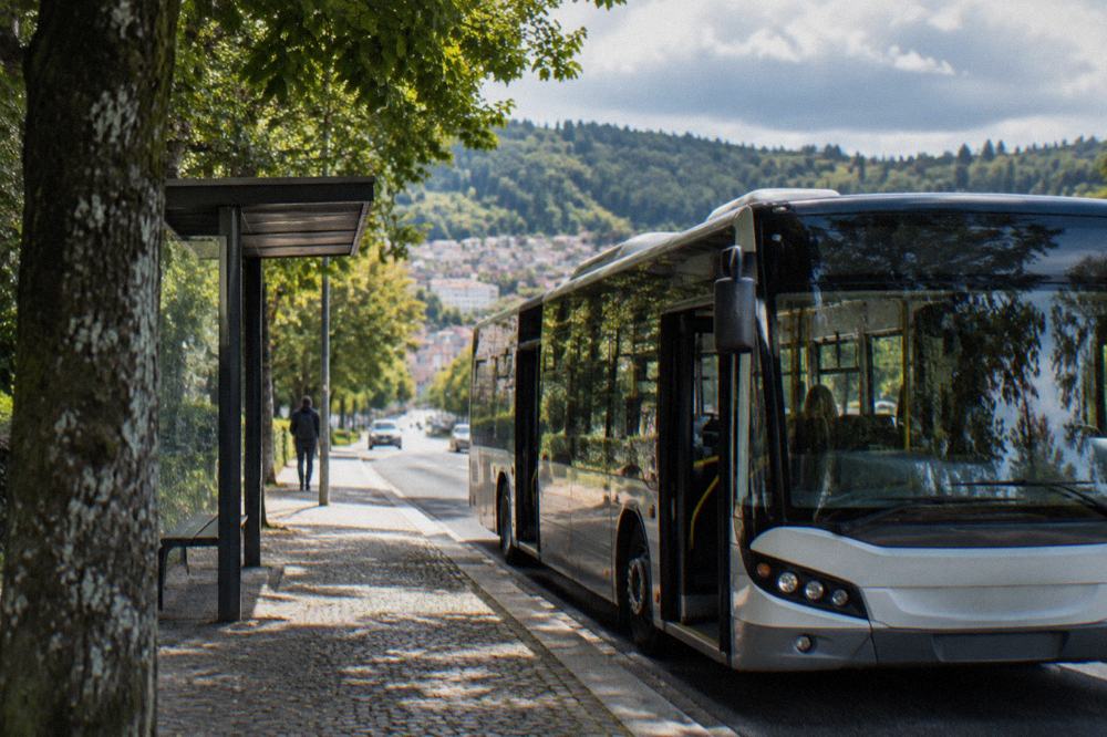 A city bus stopped at a street stop with tree-lined hills in the background