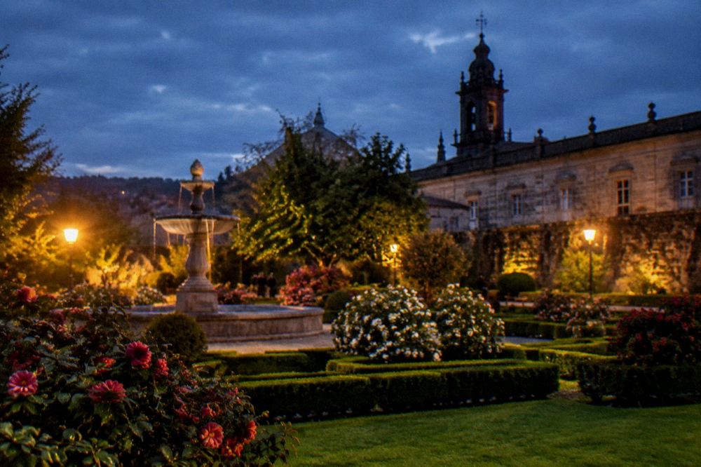 An evening view across a formal garden with lit flower beds and the dark silhouette of a church tower above