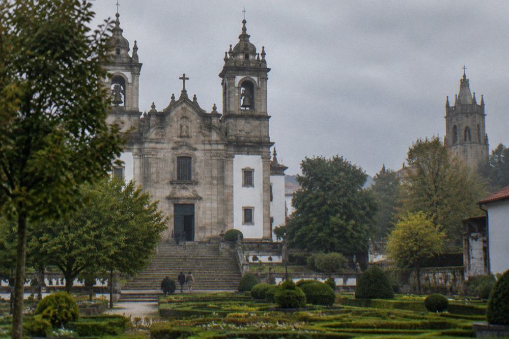 A formal garden with clipped hedges and a church tower rising behind the trees in Braga