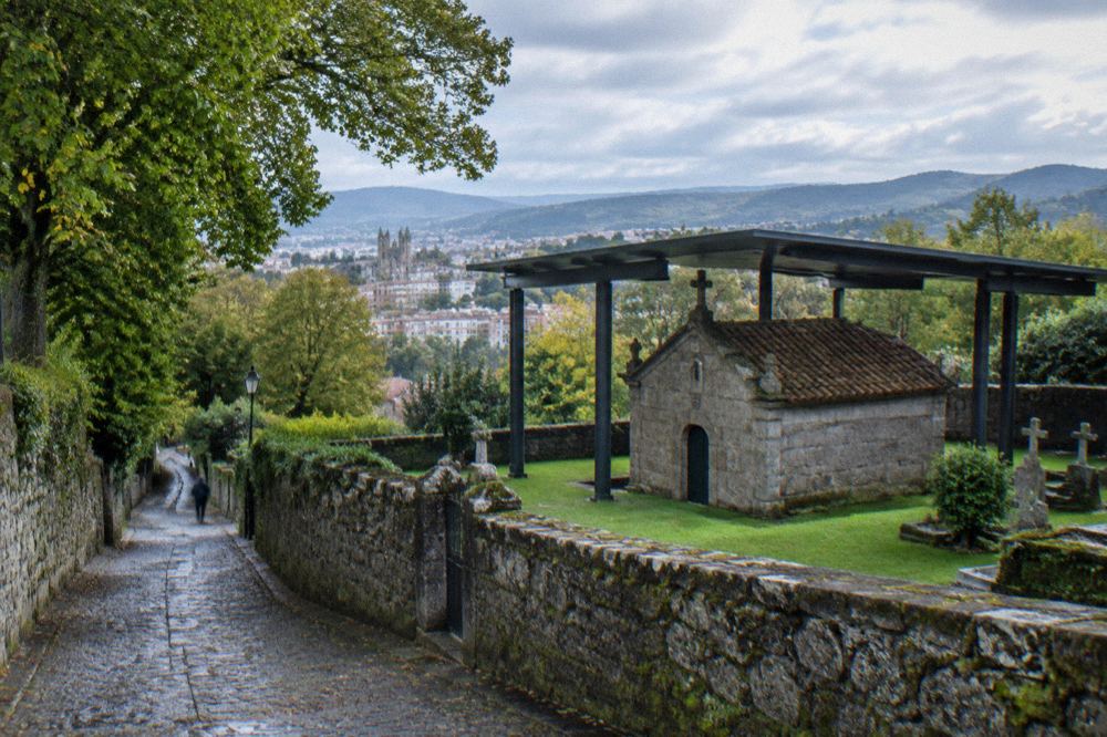 A narrow lane climbing through trees toward a small stone chapel on the edge of a city