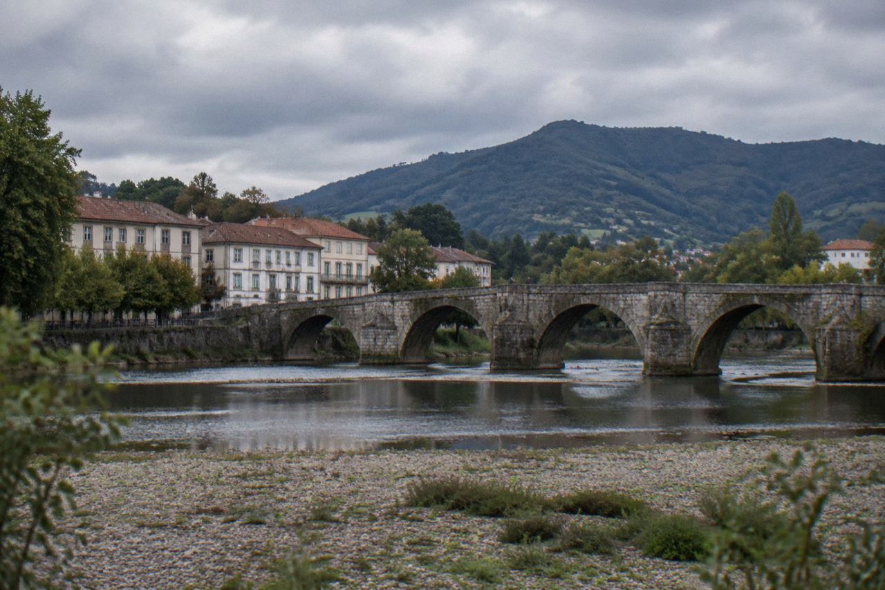 A stone bridge over a calm river with a small town and green hills behind it