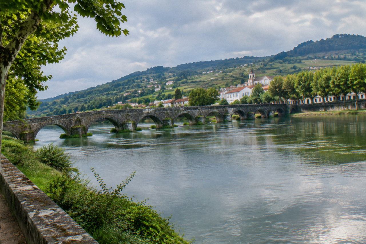 A stone bridge with arches crossing a wide green river towards a small town with trees along the banks
