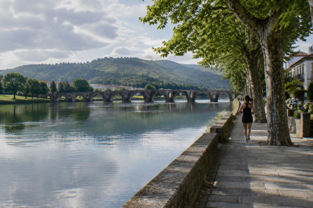 A wide slow river reflecting trees and a distant stone bridge, with green fields on the far bank