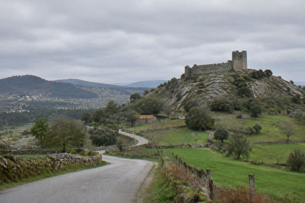 A narrow country road winding through green Minho farmland towards a rocky hill