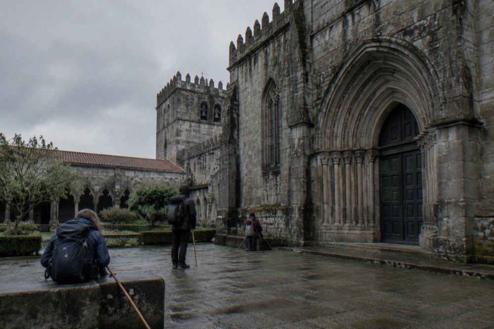Pilgrims with backpacks and walking staffs resting on the stone pavement beside a grand cathedral facade