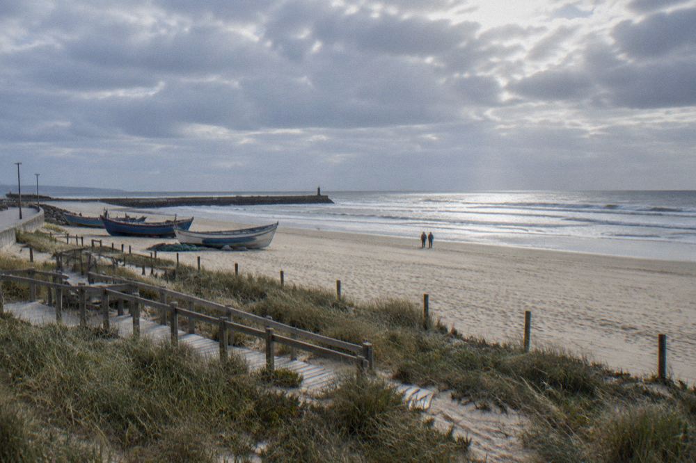 A wide sandy beach with a promenade and fishing boats near a harbour wall