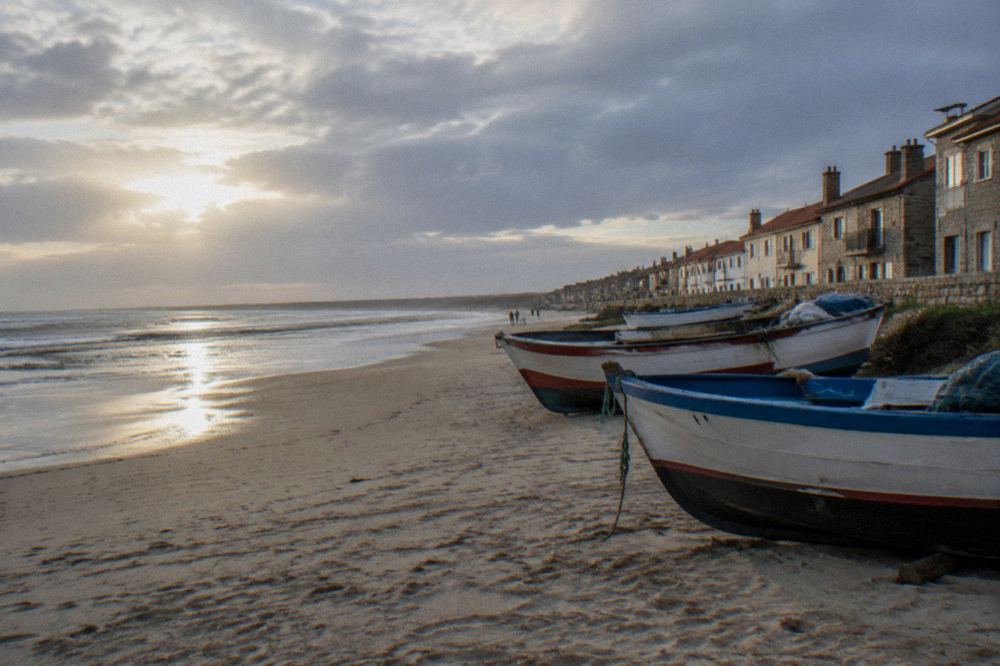 Fishing boats resting on a sandy beach beside a row of stone houses at a Portuguese seaside town