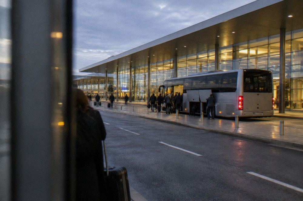 A modern airport terminal forecourt at dusk with a coach loading passengers at a kerbside stop