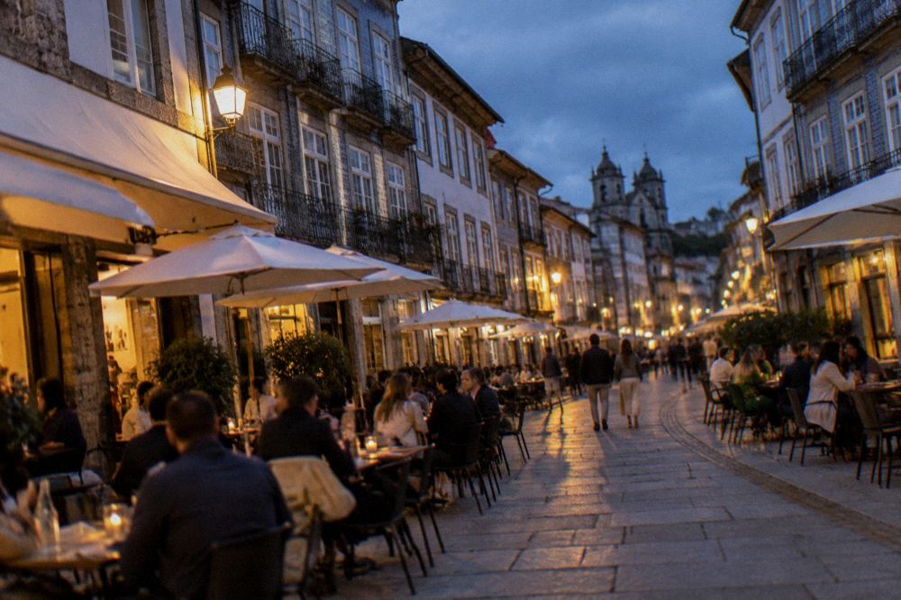 A busy terrace of small tables with people eating outdoors on a warm evening in a European square