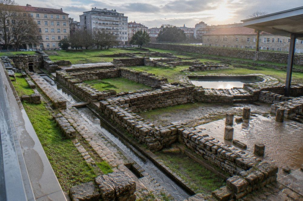 The stone foundations of an ancient Roman bath complex seen from a raised walkway