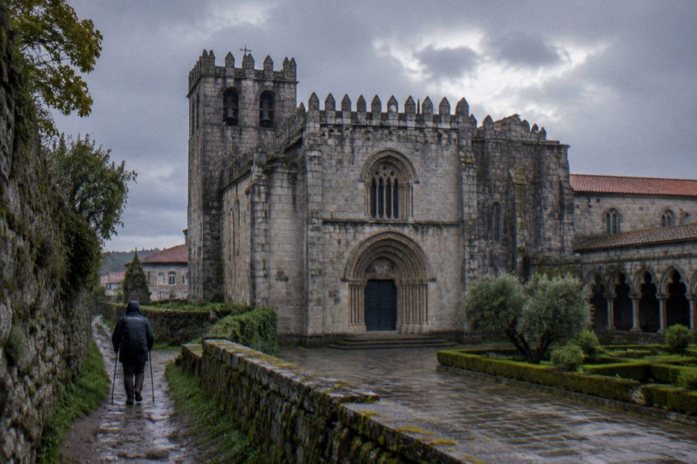 A pilgrim walking a muddy rural path between stone walls in light rain