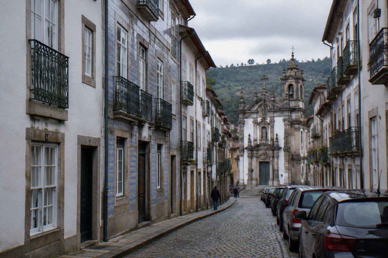 A narrow cobbled street in Braga's historic centre with parked cars lining one side and baroque buildings above