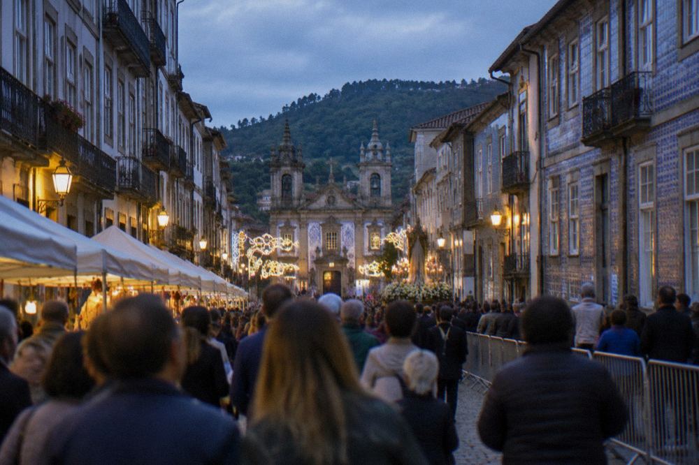 A crowded square in Braga during a festival evening with lights, stalls and a procession route barrier in place
