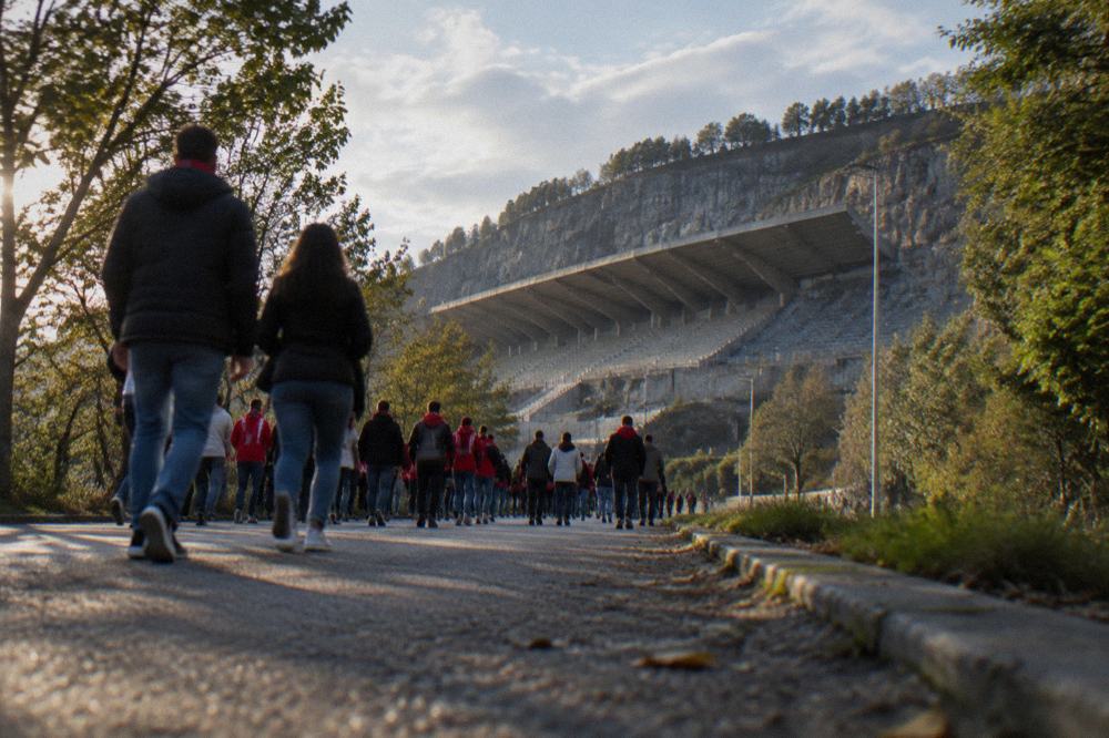 Crowd of supporters walking along a tree-lined road toward a stadium before kickoff