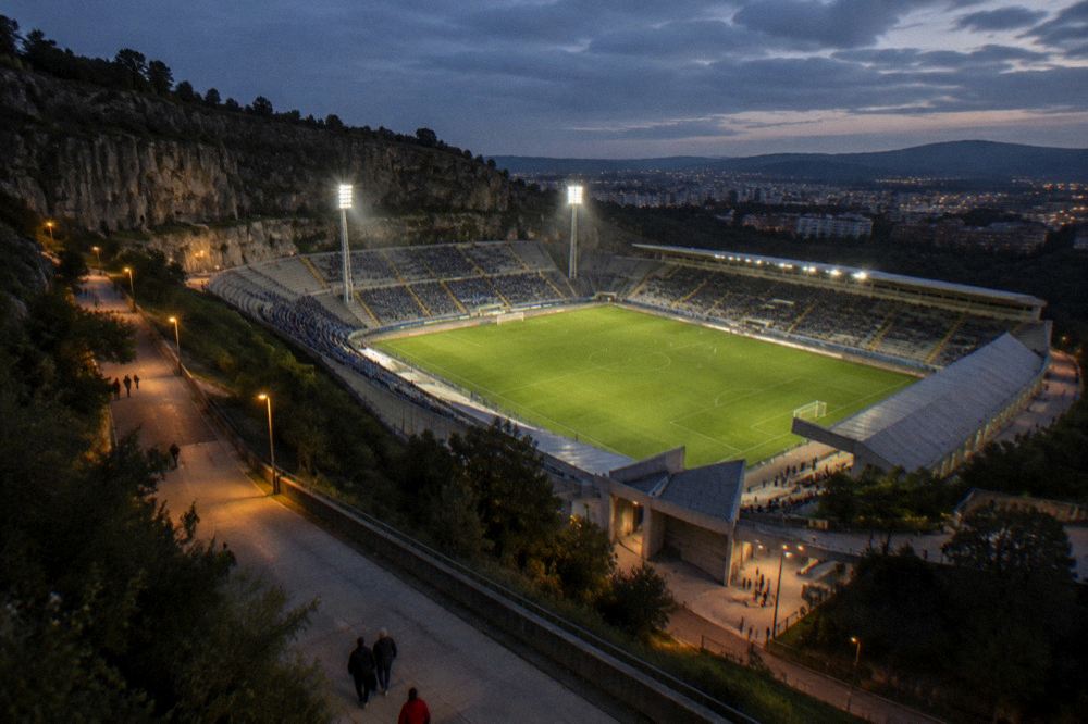 Stadium floodlights glowing over a pitch at dusk with the first spectators in the stands