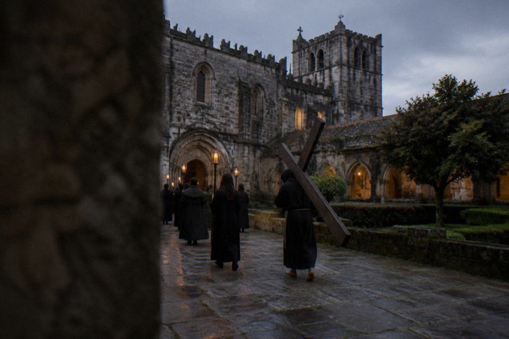 A barefoot penitent in a dark robe carrying a heavy wooden cross surrounded by candle bearers at night