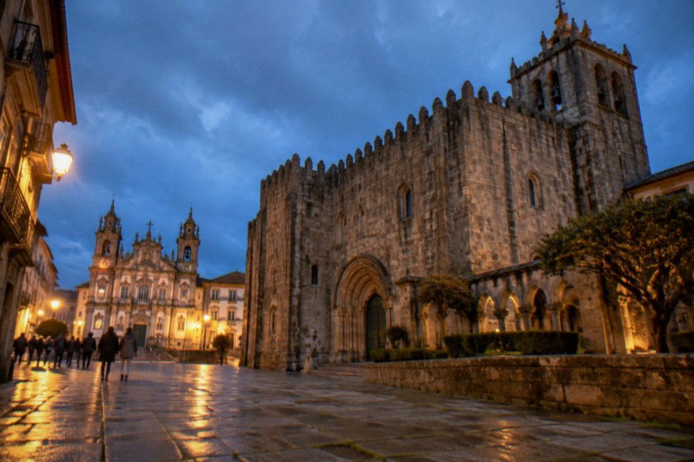 A illuminated historic city square at dusk with a baroque church facade and people walking