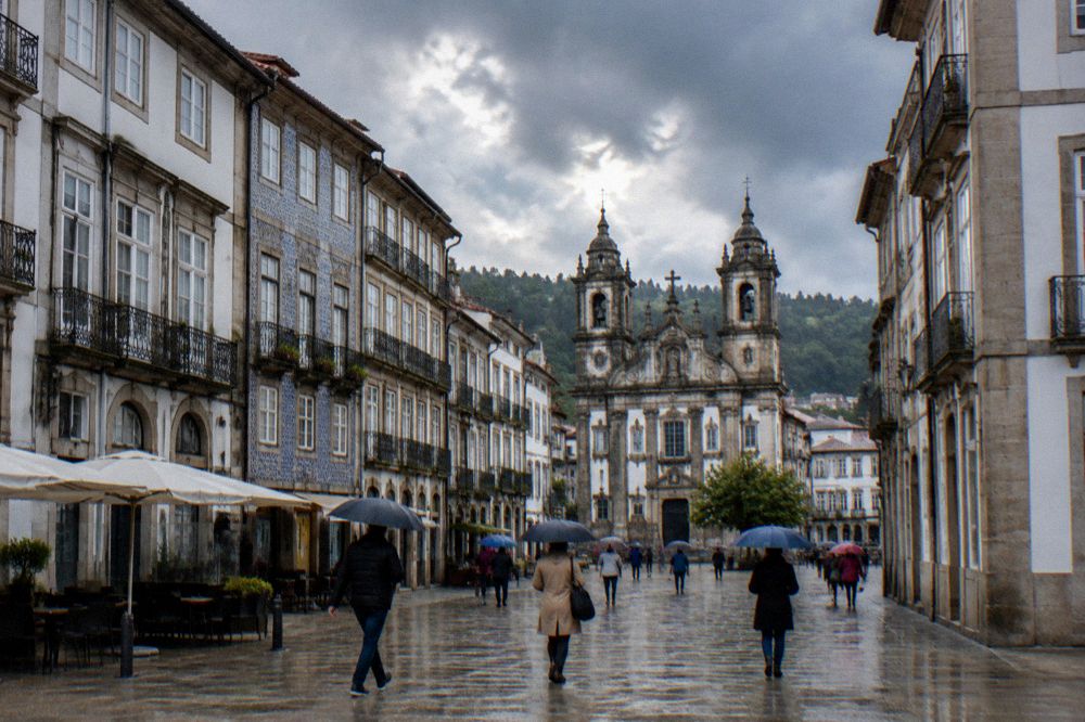 Pedestrians with umbrellas crossing a wet granite square in central Braga